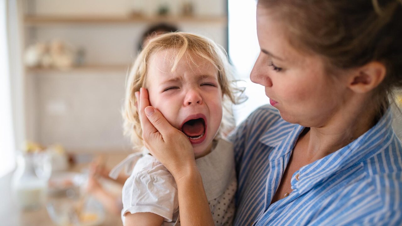 A mother holding a crying toddler daughter indoors in kitchen when cooking.