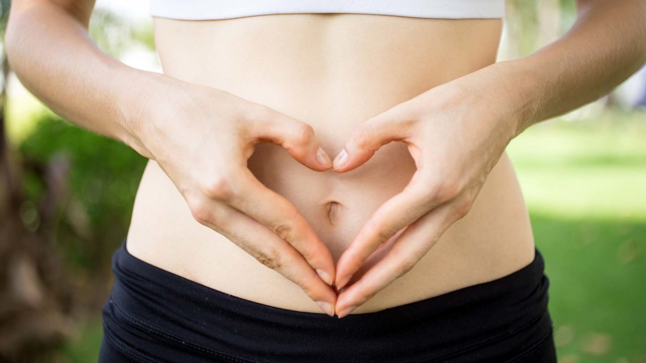 Close-up of female hands shaping heart on belly outdoors. Fitness and dieting concept